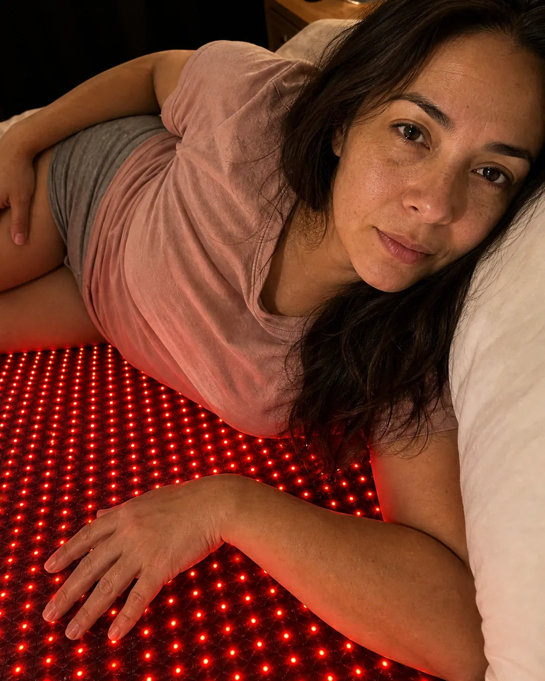 Woman lying on her side on the EMBRA red light mat on her bed at night, bedside lamp on