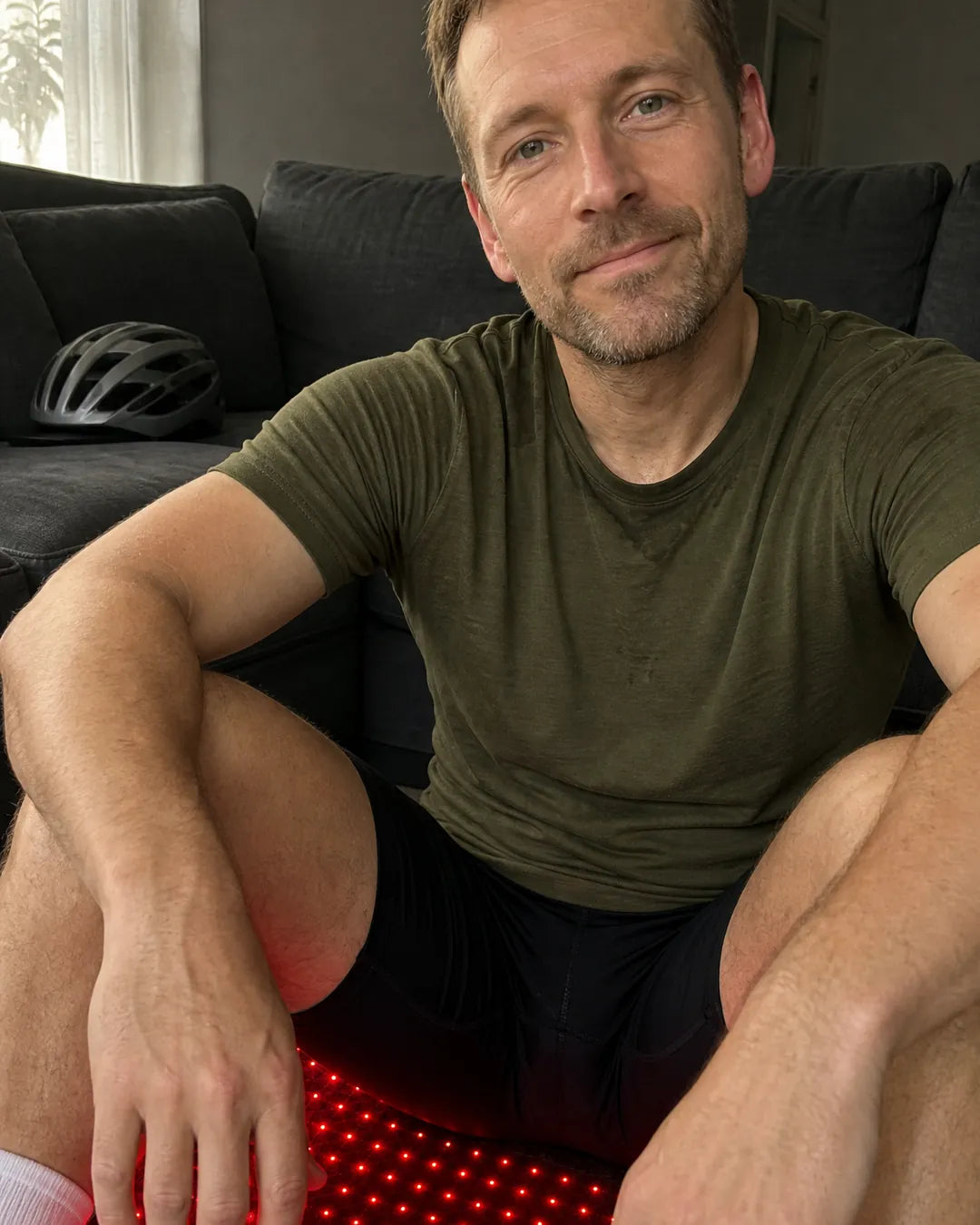 Man in cycling shorts sitting on the EMBRA red light mat against the sofa, bike helmet behind him